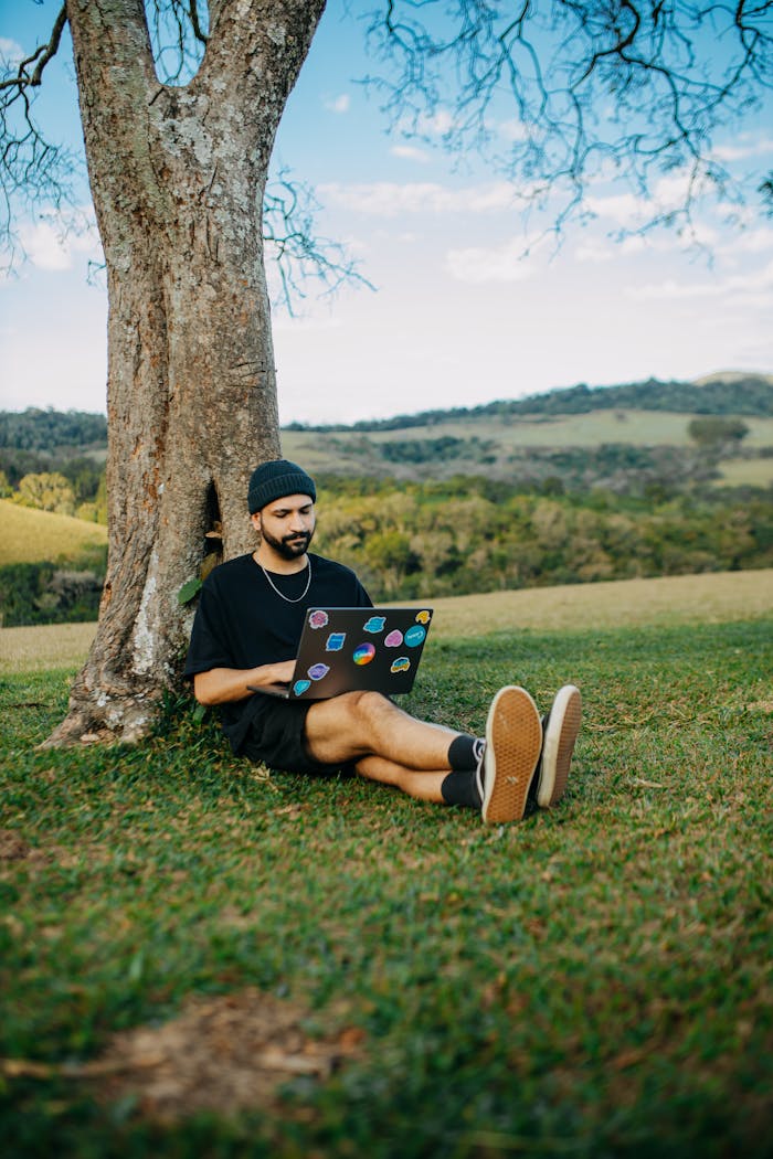 Man sitting under a tree with a laptop, surrounded by lush greenery and scenic hills.