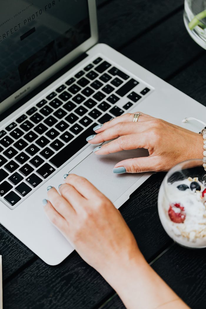 Close-up of woman's hands on laptop and a bowl of yogurt with berries on a black table.