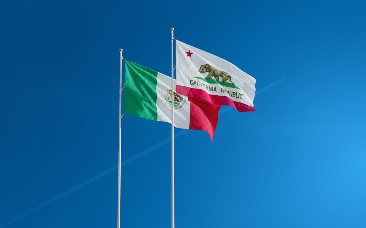 Mexican and Californian flags waving on poles against a clear blue sky.