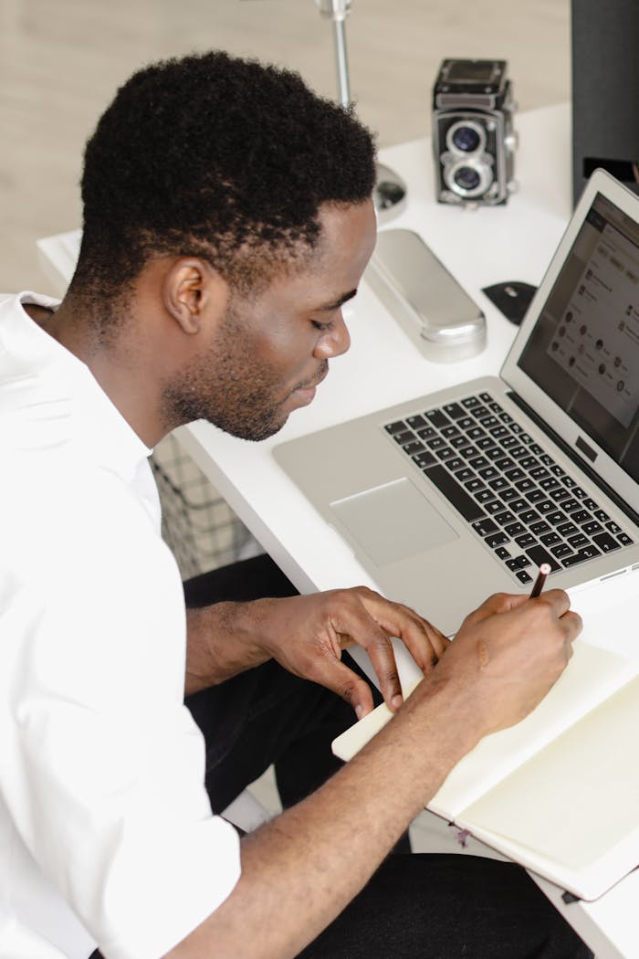 Man in office setting, actively engaged with laptop and notebook, taking notes.